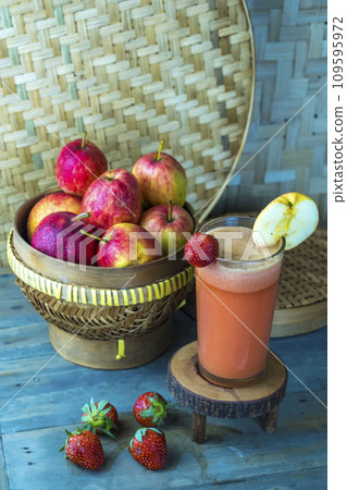 A basket of red apples and strawberries next to a glass of apple and strawberry juice on a wooden table. 109595972