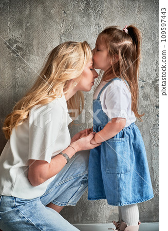 Side view of cute child kissing mother while standing against the wall at home. Blonde woman holding daughter hands while kid placing kiss on mother forehead. Family and motherhood concept. 109597443