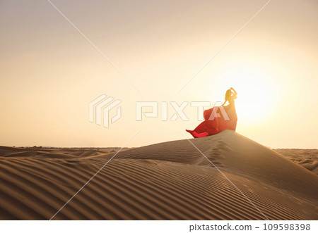 Desert adventure. Young arabian Woman in red silk dress in sands dunes of UAE desert at sunset, fantastic view. The Dubai Desert Conservation Reserve, United Arab Emirates. 109598398