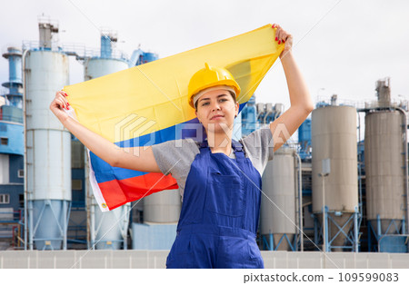 Positive young female worker in helmet waving national flag of Colombia while standing in front of big tanks at refinery factory on sunny summer day 109599083