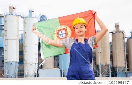 Young female engineer in helmet waving state flag of Portugal while standing in front of big tanks at chemical plant 109599157