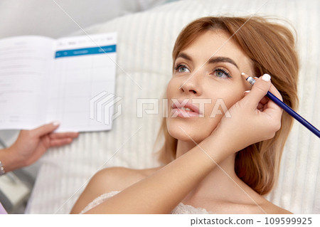 cosmetologist marking skin of caucasian woman with white pencil. facial marking for aesthetic procedure. cosmetologist marking skin of caucasian woman with white pencil. facial marking for aesthetic procedure. 109599925