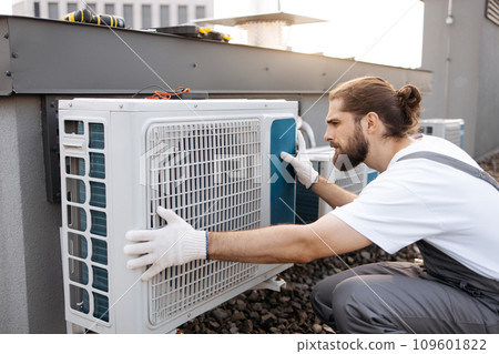 Man construction crouching and examining conditioner on roof 109601822