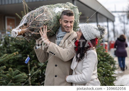 A young man carrying a wrapped Christmas tree bought at the market together with his wife. The happy couple bought a Christmas tree 109602056