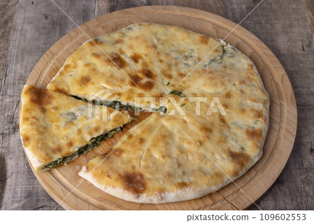 Pie with spinach, herbs and suluguni cheese on a wooden board. Whole pie with cut off piece. Traditional round flat pie with filling. Homemade pastries of Caucasian and Balkarian cuisine. Pie with spinach, herbs and suluguni cheese on a wooden board. Whole pie with cut off piece. Traditional round flat pie with filling. Homemade pastries of Caucasian and Balkarian cuisine. 109602553
