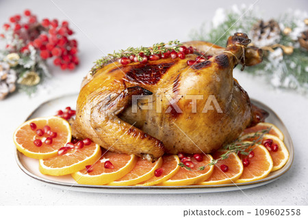 Chicken baked whole to appetizing crispy crust with pomegranate and red orange. Traditional New Year and Christmas dish. In the background are Christmas decorations. Close-up, selective focus. 109602558