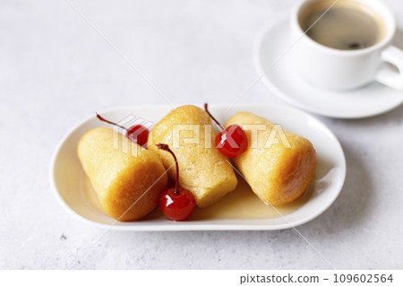 Neapolitan Rum baba (or baba au rum) on a white plate with a cocktail cherry and a cup of coffee. Small yeast cakes soaked in rum syrup on a gray background. Traditional Italian pastry. Selective focu Neapolitan Rum baba (or baba au rum) on a white plate with a cocktail cherry and a cup of coffee. Small yeast cakes soaked in rum syrup on a gray background. Traditional Italian pastry. Selective focu 109602564