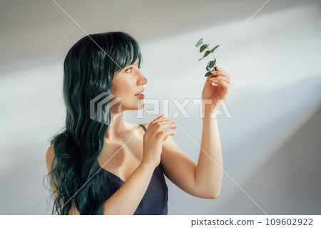 Portrait of Adorable tender young woman with long green color hair holding eucalyptus branch in hand in ray of sun from window on light background. Concept of beauty, art, fashion. Mermaid style. Portrait of Adorable tender young woman with long green color hair holding eucalyptus branch in hand in ray of sun from window on light background. Concept of beauty, art, fashion. Mermaid style. 109602922
