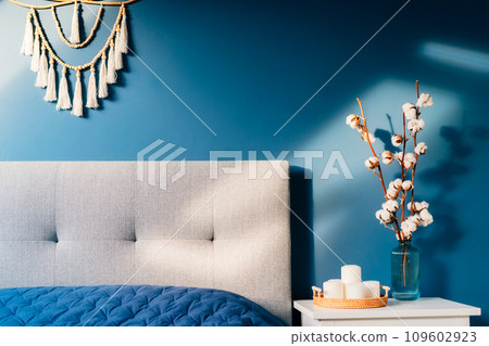 Stylish modern bedroom in dark colors. Cozy interior with navy blue walls, home decor. Bed with gray fabric headboard, blue blanket, bedside table, vase with natural cotton flowers, candles on tray Stylish modern bedroom in dark colors. Cozy interior with navy blue walls, home decor. Bed with gray fabric headboard, blue blanket, bedside table, vase with natural cotton flowers, candles on tray 109602923