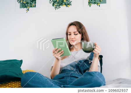 Young woman reading e-book on tablet reader in bed. Female relaxing with hot drink while reading an electronic book in cozy room. Mental health recovery, Personal growth. Hobby, Leisure time alone. Young woman reading e-book on tablet reader in bed. Female relaxing with hot drink while reading an electronic book in cozy room. Mental health recovery, Personal growth. Hobby, Leisure time alone. 109602937