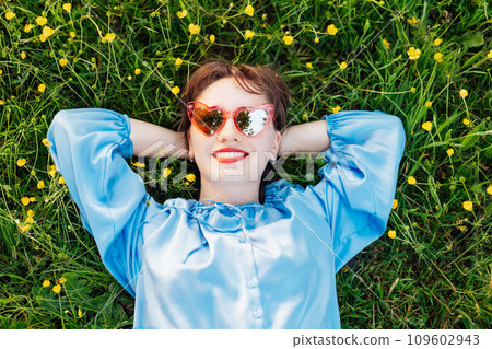 Top view happy young woman in blue blouse lays on a green grass lawn with yellow flowers, looks up and smiles. Business woman enjoy the moment, relaxing outdoors. Spring or summer season concept. Top view happy young woman in blue blouse lays on a green grass lawn with yellow flowers, looks up and smiles. Business woman enjoy the moment, relaxing outdoors. Spring or summer season concept. 109602943