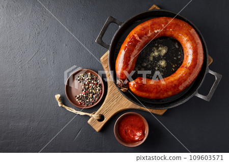 Fried pork sausage on a black cast iron pan on a wooden board and peppercorns with tomato sauce on a black stone table. Top view with copy space Fried pork sausage on a black cast iron pan on a wooden board and peppercorns with tomato sauce on a black stone table. Top view with copy space 109603571
