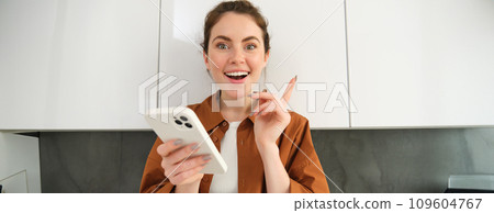 Portrait of excited young woman has an idea, holding smartphone and pointing finger up, standing in the kitchen at home 109604767