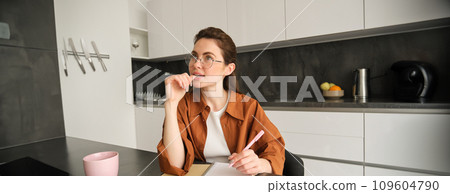 Young creative woman, writer making notes, holding pen, looking aside with thinking face, sitting in kitchen at home, doing homework Young creative woman, writer making notes, holding pen, looking aside with thinking face, sitting in kitchen at home, doing homework 109604790