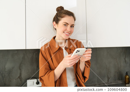 Portrait of young smiling woman with mobile phone, standing in the kitchen, looking happy and relaxed at camera Portrait of young smiling woman with mobile phone, standing in the kitchen, looking happy and relaxed at camera 109604800
