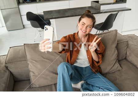 Portrait of beautiful smiling woman, takes selfie at home, posing on sofa, holds smartphone with extended hand Portrait of beautiful smiling woman, takes selfie at home, posing on sofa, holds smartphone with extended hand 109604802