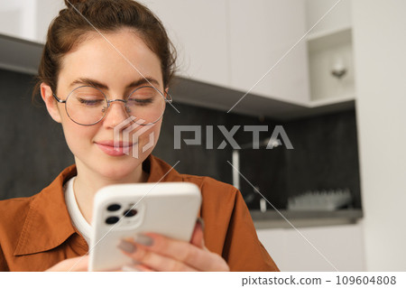 Portrait of young brunette woman in glasses, sitting at home with mobile phone, using her smartphone and smiling, reading message 109604808