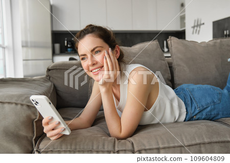 Portrait of cute, happy young woman lying on couch, holding mobile phone, using smartphone while relaxing at home in living room, concept of lifestyle and people leisure 109604809