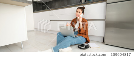 Young woman casually sits on floor with laptop, makes a phone call, talking on telephone 109604817