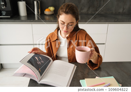 Distance learning. Young woman at home, student studying at home and drinking tea, reading her work book, revising for exam 109604822