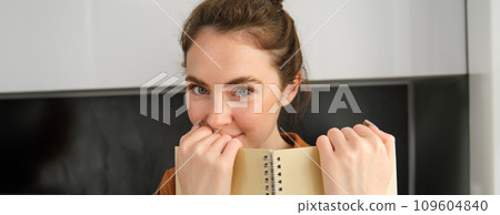 Close up portrait of cute girlfriend, giggle, reading notebook, holding planner and smiling at camera Close up portrait of cute girlfriend, giggle, reading notebook, holding planner and smiling at camera 109604840
