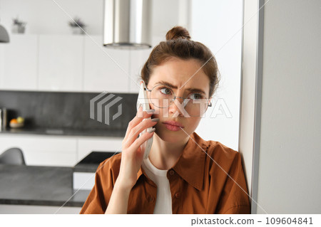 Portrait of woman with disappointed face, standing at home, answers phone call with concerned face, has difficult telephone conversation Portrait of woman with disappointed face, standing at home, answers phone call with concerned face, has difficult telephone conversation 109604841