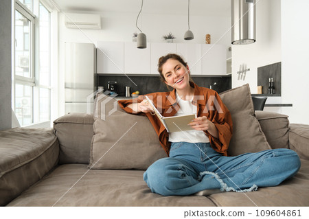 Young woman sits at home on sofa, reading her notes, holding notebook, studying for exam, student revising in her living room 109604861