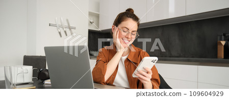Portrait of young woman freelancer, working from home, saleswoman doing trading from her kitchen, set up workplace in flat, sitting near laptop, using smartphone 109604929