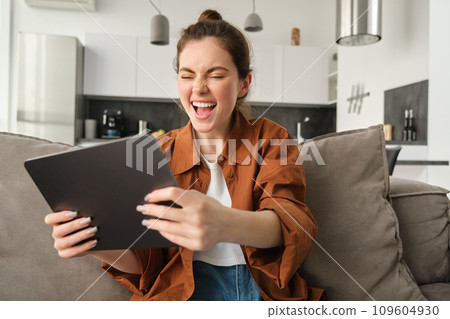 Portrait of excited young woman with digital tablet, sitting on couch, laughing and smiling, winning on video game, spending time at home 109604930