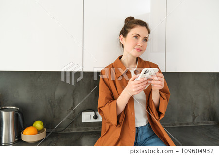 Image of young woman with smartphone, looking with thoughtful expression, buying online using mobile app, orders food delivery with phone application 109604932