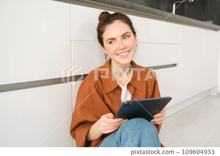 Portrait of beautiful, carefree young woman, sitting on kitchen floor with digital tablet, working remote from home, using device to check her mail. Girl watching something on her device 109604933