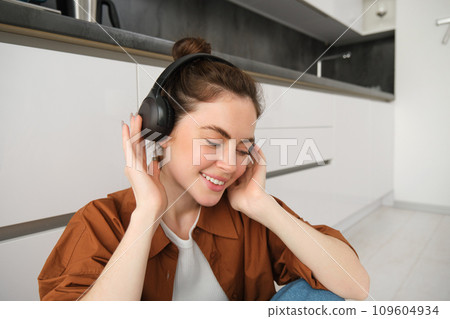 Cute brunette woman on kitchen floor, wears headphones, listens music and smiles, enjoys sound quality 109604934