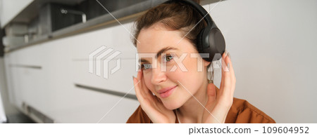 Image of modern woman, beautiful girl enjoys listening to music, has black wireless headphones on, sits on floor and rests 109604952