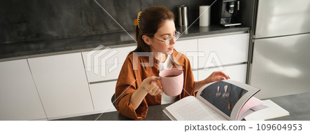 Portrait of beautiful young woman in kitchen, sitting with a book, flipping pages, reading and drinking coffee Portrait of beautiful young woman in kitchen, sitting with a book, flipping pages, reading and drinking coffee 109604953