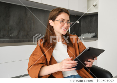 Portrait of young student, woman studying at home, working remotely, setup workplace in her kitchen, sitting on chair with digital tablet, reading in glasses 109605092