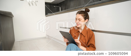 Lifestyle shot of young woman, student using her digital tablet, laughing and smiling, sitting on kitchen floor at home Lifestyle shot of young woman, student using her digital tablet, laughing and smiling, sitting on kitchen floor at home 109605094
