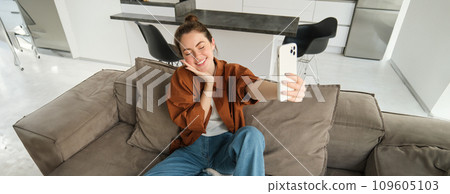 Portrait of beautiful smiling woman, takes selfie at home, posing on sofa, holds smartphone with extended hand 109605103