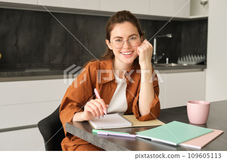 Portrait of young smiling woman, tutor preparing notes for lesson at home, writing in notebook and looking happy, student studying in kitchen Portrait of young smiling woman, tutor preparing notes for lesson at home, writing in notebook and looking happy, student studying in kitchen 109605113