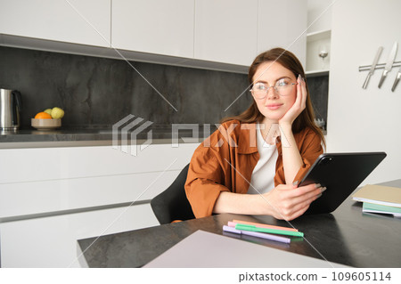 Portrait of young student, woman studying at home, working remotely, setup workplace in her kitchen, sitting on chair with digital tablet, reading in glasses Portrait of young student, woman studying at home, working remotely, setup workplace in her kitchen, sitting on chair with digital tablet, reading in glasses 109605114
