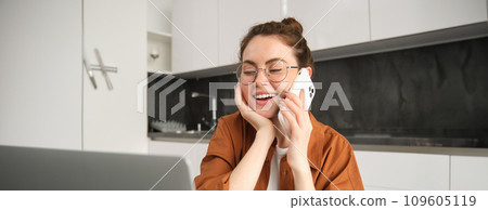 Portrait of beautiful young woman managing her own business from home, freelancer making phone call, sitting in kitchen with laptop and smiling Portrait of beautiful young woman managing her own business from home, freelancer making phone call, sitting in kitchen with laptop and smiling 109605119