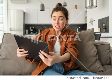 Portrait of anxious, stressed young woman looking scared at digital tablet, staring at screen, sitting on couch with gadget in living room 109605152