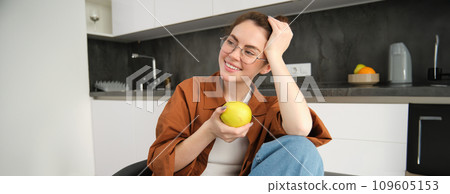 Beautiful young woman in glasses, sitting on chair in the kitchen, eating green apple and smiling 109605153