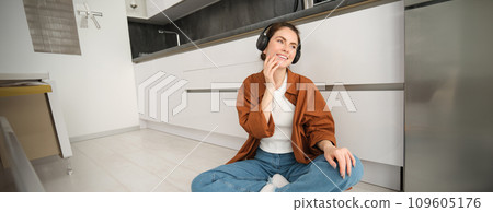 Cute brunette woman on kitchen floor, wears headphones, listens music and smiles, enjoys sound quality Cute brunette woman on kitchen floor, wears headphones, listens music and smiles, enjoys sound quality 109605176