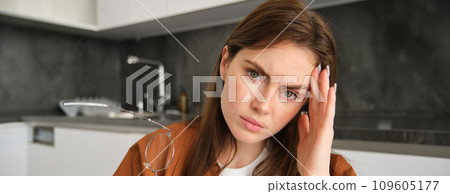 Close up portrait of tired young woman, wife sitting in kitchen and touching head, has headache, massaging temple and frowning from painful migraine 109605177