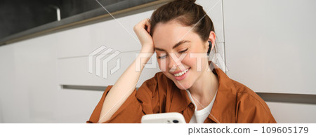 Portrait of happy young beautiful woman, listening music in wireless earphones, using smartphone, watching videos on mobile phone with headphones on 109605179