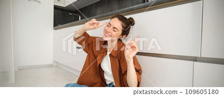 Portrait of young carefree woman sits on floor at home, dances while listens to music in wireless headphones, singing along favourite song 109605180