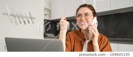 Portrait of young woman at home, sitting in kitchen with laptop, talking on mobile phone, making a call, working in her flat 109605195