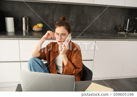Portrait of busy young self-employed woman, working from home, has office in her flat, sitting near laptop, answers phone call, talking with serious, concerned face expression Portrait of busy young self-employed woman, working from home, has office in her flat, sitting near laptop, answers phone call, talking with serious, concerned face expression 109605196