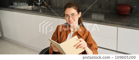 Portrait of young self-employed freelance tutor, sitting in kitchen at home, holding notebook with study notes, revising for university 109605200