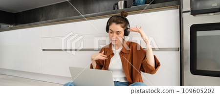 Carefree young woman sits on kitchen floor with laptop, freelancing, digital nomad working from home on computer, listening to music 109605202